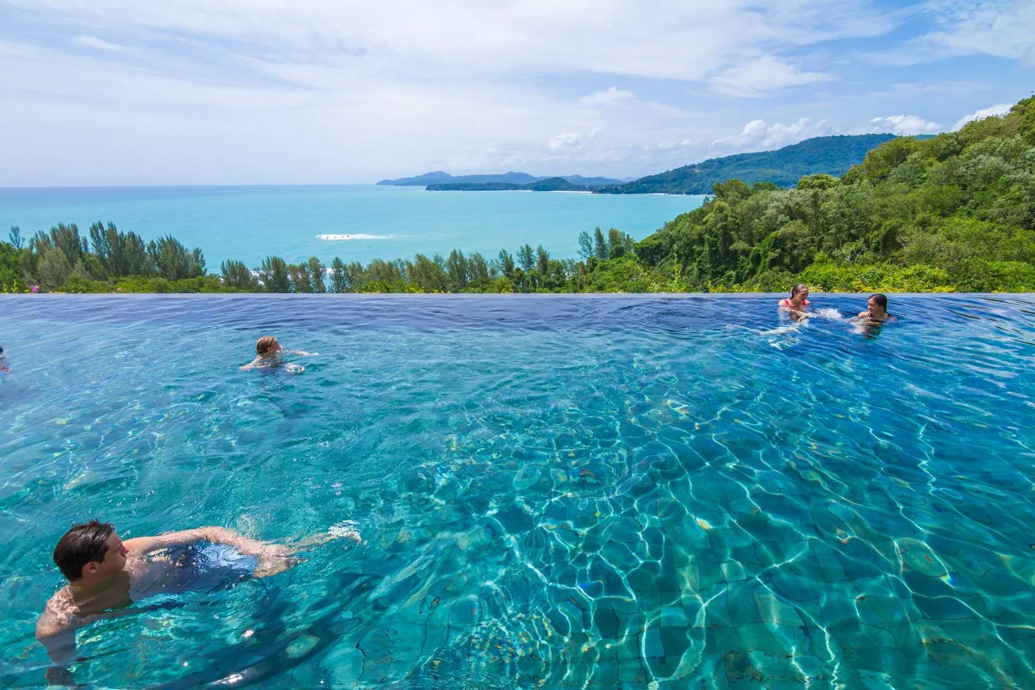 An infinity pool overlooking the sea with two people swimming, forested coastline in the distance