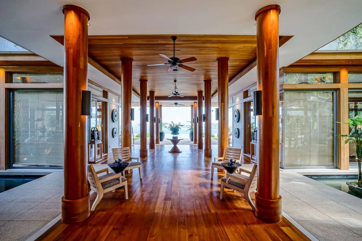 A long covered walkway with timber columns and wood flooring leading to a seating area, with a sea view at the far end