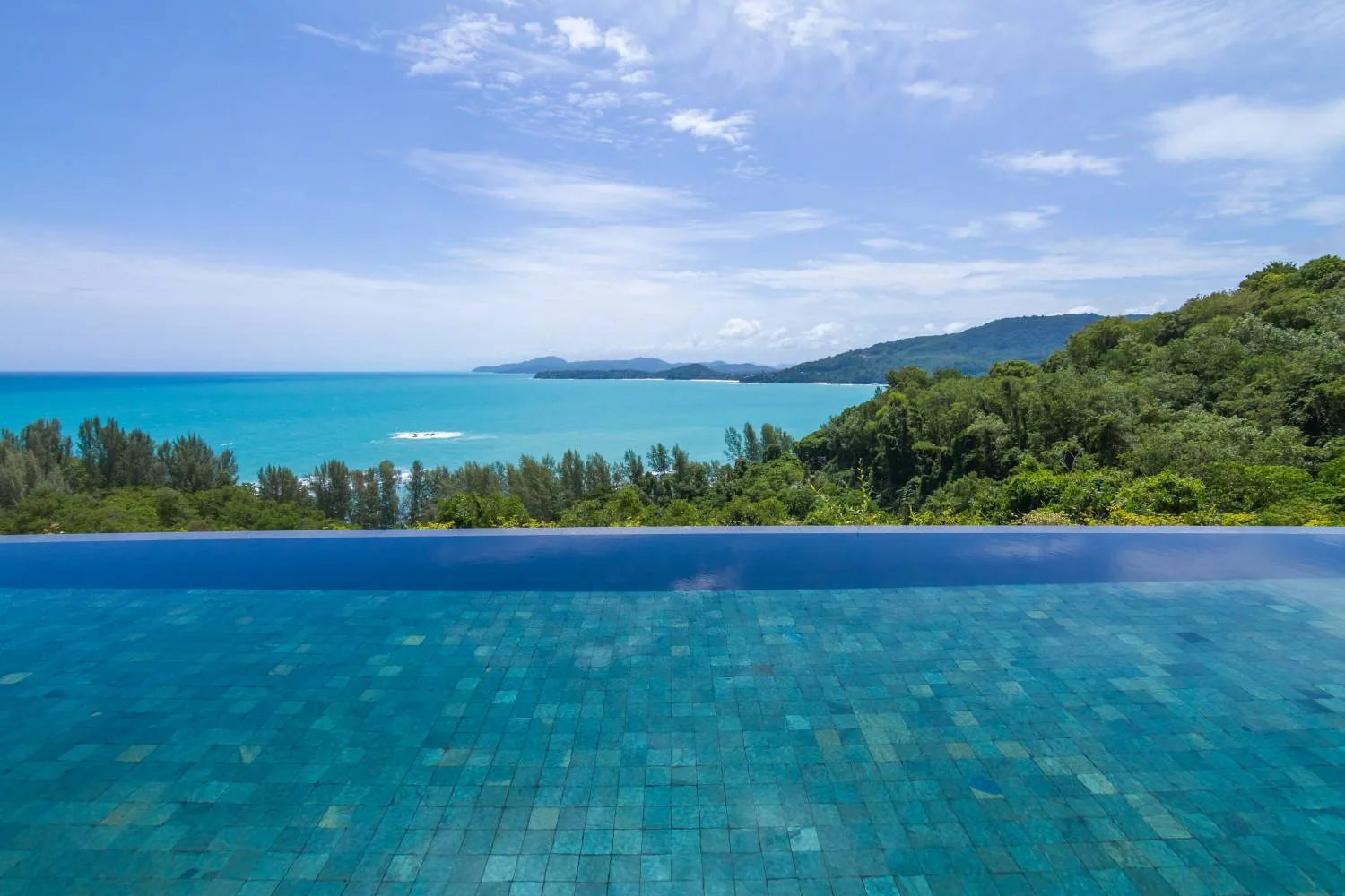 The edge of an infinity pool with teal mosaic tiles, looking out over the sea and forested hills