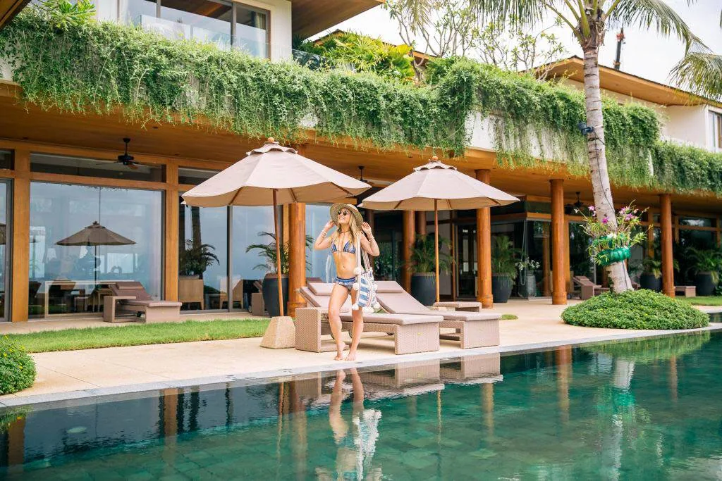 A pool deck with sun loungers and umbrellas; a person stands at the poolside with the villa building behind