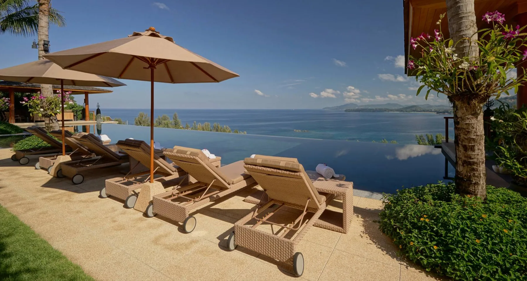 Sun loungers under a shade umbrella on a terrace overlooking the sea, with palm trees in view