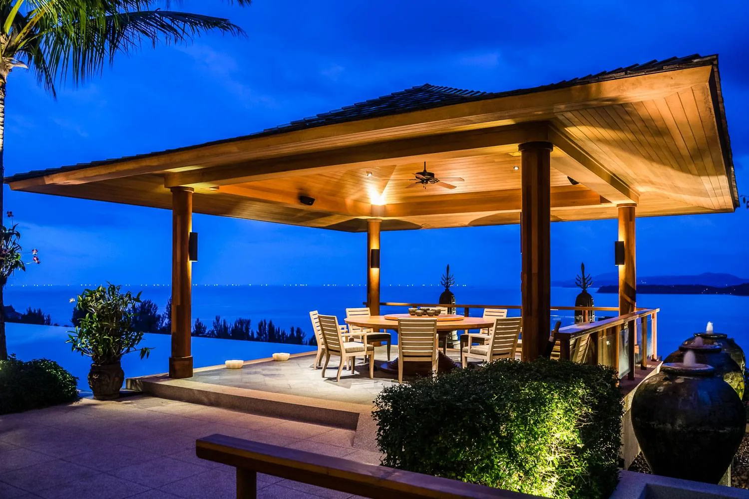 An open sided timber pavilion set up for dining, lit at dusk with a sea view beyond
